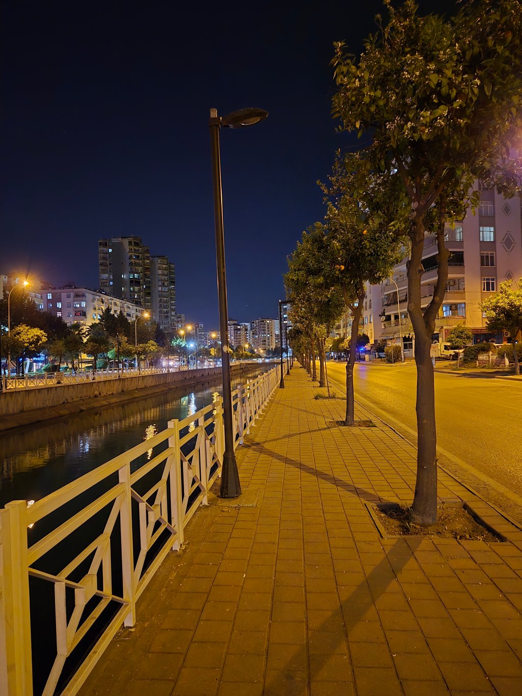 Gece saatlerinde, bir şehir yürüyüş yolunun görüntüsü. Solda ağaçlar ve aydınlatma direkleri, sağda bir su kanalı ve binalar yer almakta. Şehrin gece atmosferi belirgin.