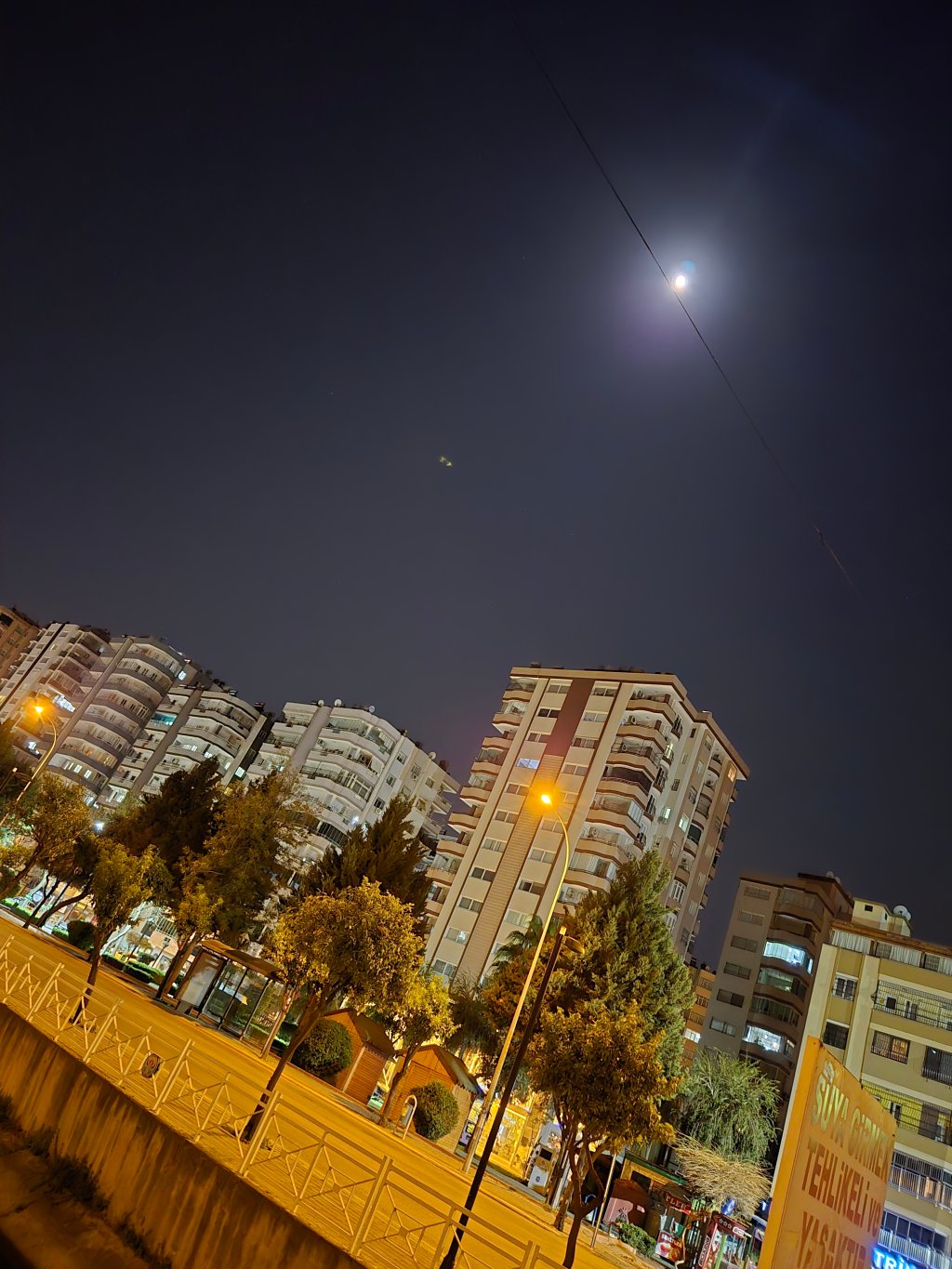 Gece çekimi, binaların aydınlatmalarının ve dolunayın net bir şekilde görüldüğü bir kentsel manzara.