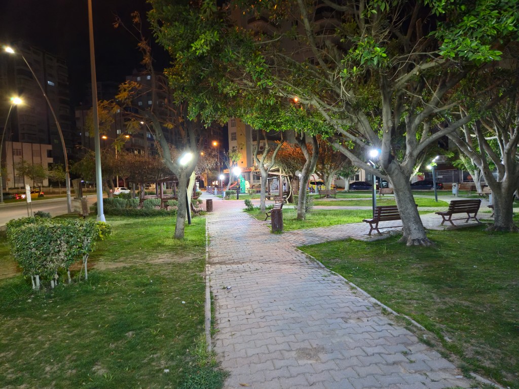 Gece çekimi yapılan bir parkın görünümü, yeşil ağaçlar ve banklarla dolu bir yürüyüş yolu, sokak lambalarının aydınlattığı alan.