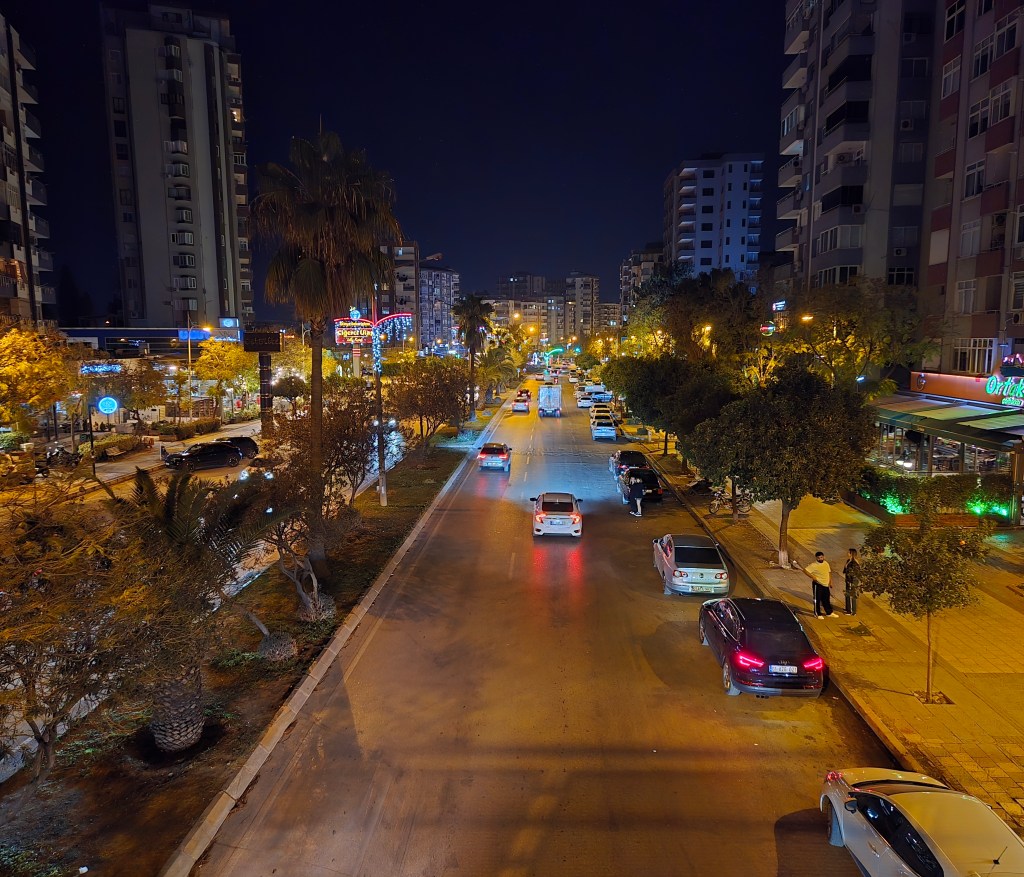 Gece çekimi, aydınlatılmış bir cadde, araçlar ve palmiyelerle çevrili bir şehir manzarası.