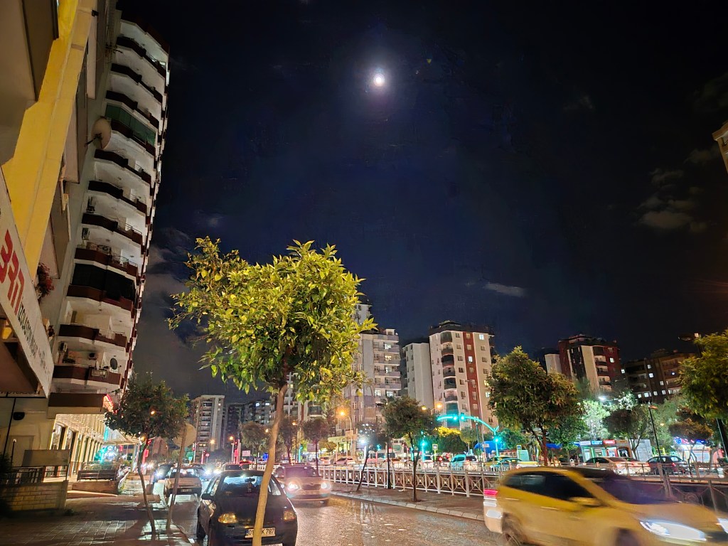 Gece vakti çekilmiş bir fotoğraf, sokaklarda aydınlatmaların parladığı ve bir ağacın ön planda yer aldığı bir manzara.